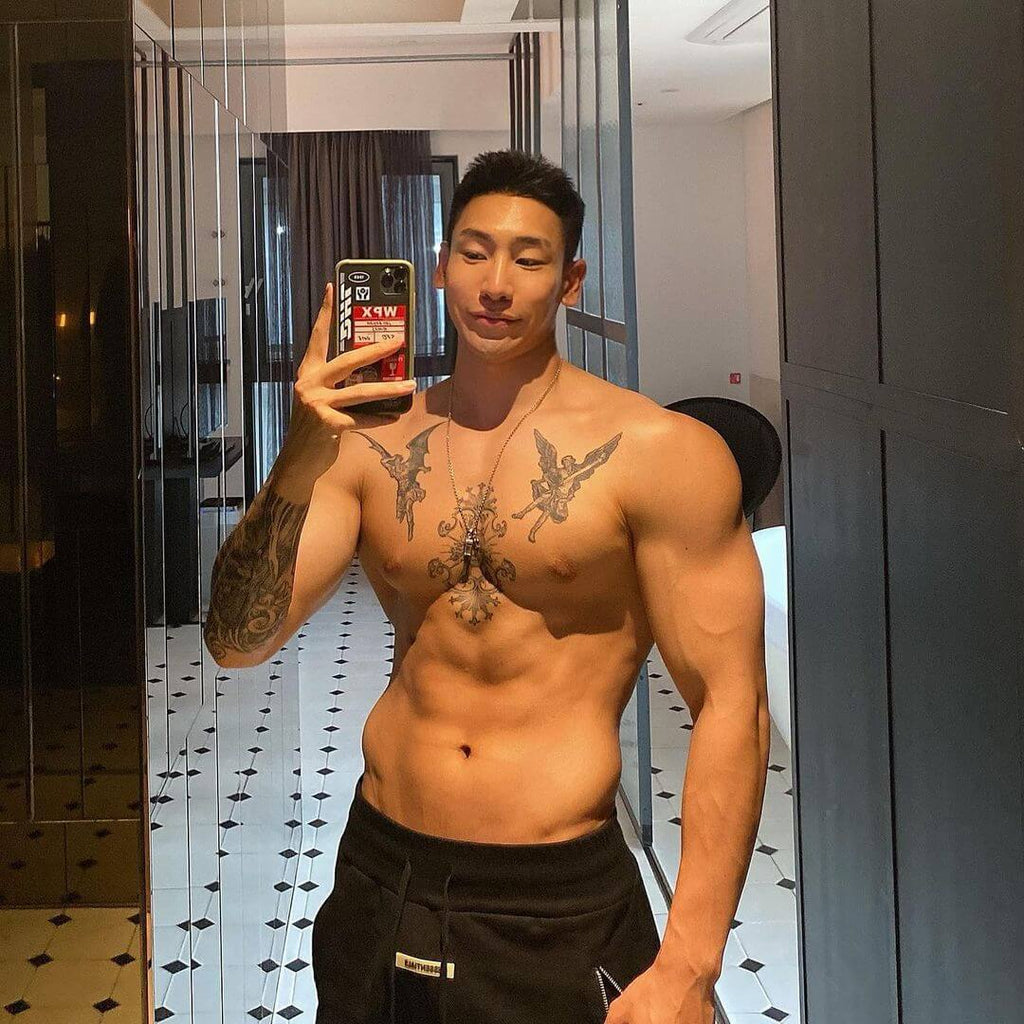 A man showcasing his muscular physique in front of a mirror, wearing fitness shorts and holding a phone.
