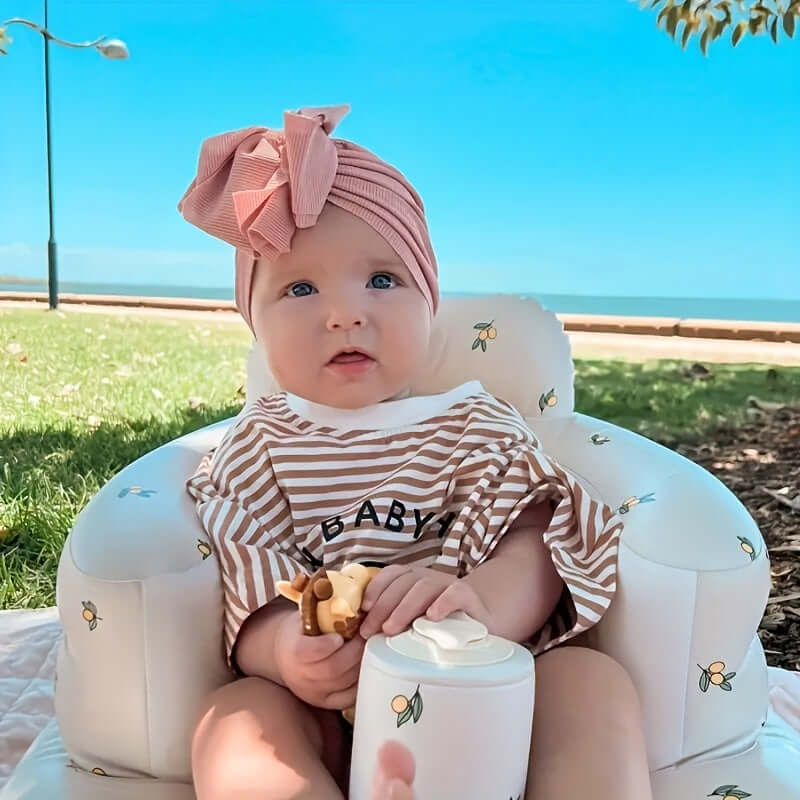 A baby sitting in The Ultimate Learning Chair For Babies, wearing a striped outfit and a cute bow.